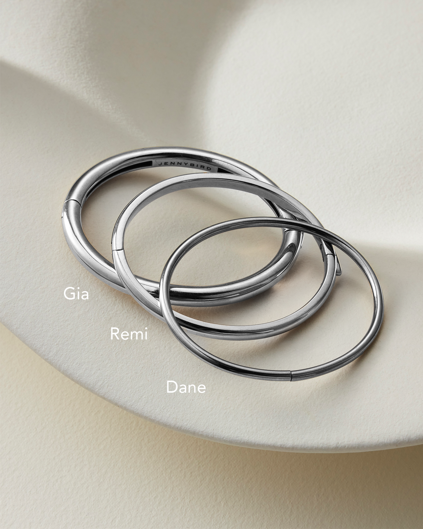 Three Silver Bangle Bracelets labeled Gia Remi and Dane laid out on a beige background for size comparison